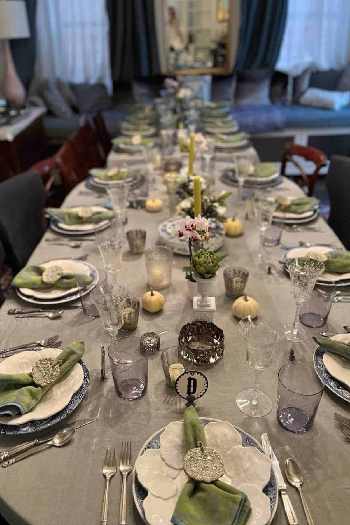 A dining table set for a formal meal with plates, glasses, cutlery, green napkins, candles, decorative pumpkins, and small floral centerpieces.