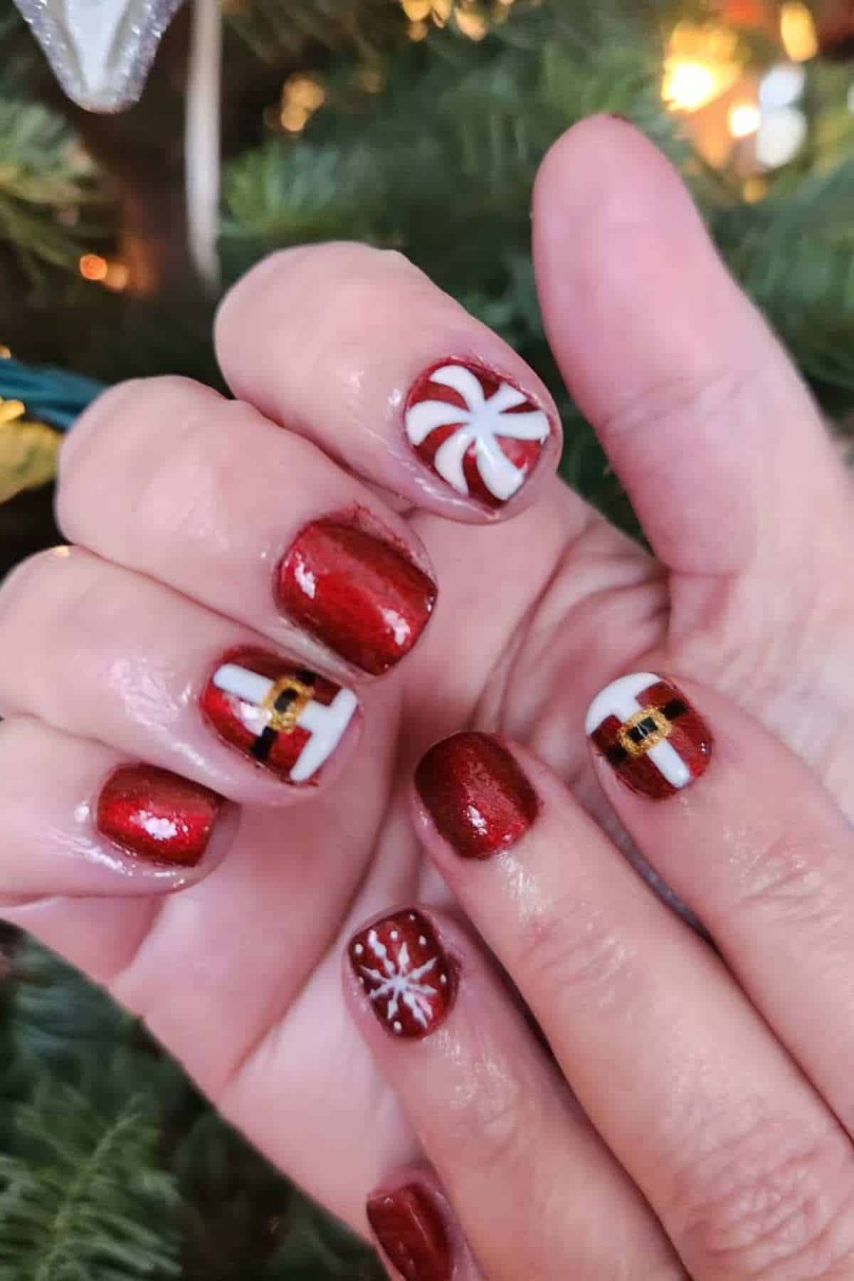 A hand with red, glossy Christmas-themed nail art featuring designs of a candy cane, snowflake, and Santa suit, held in front of a decorated Christmas tree.