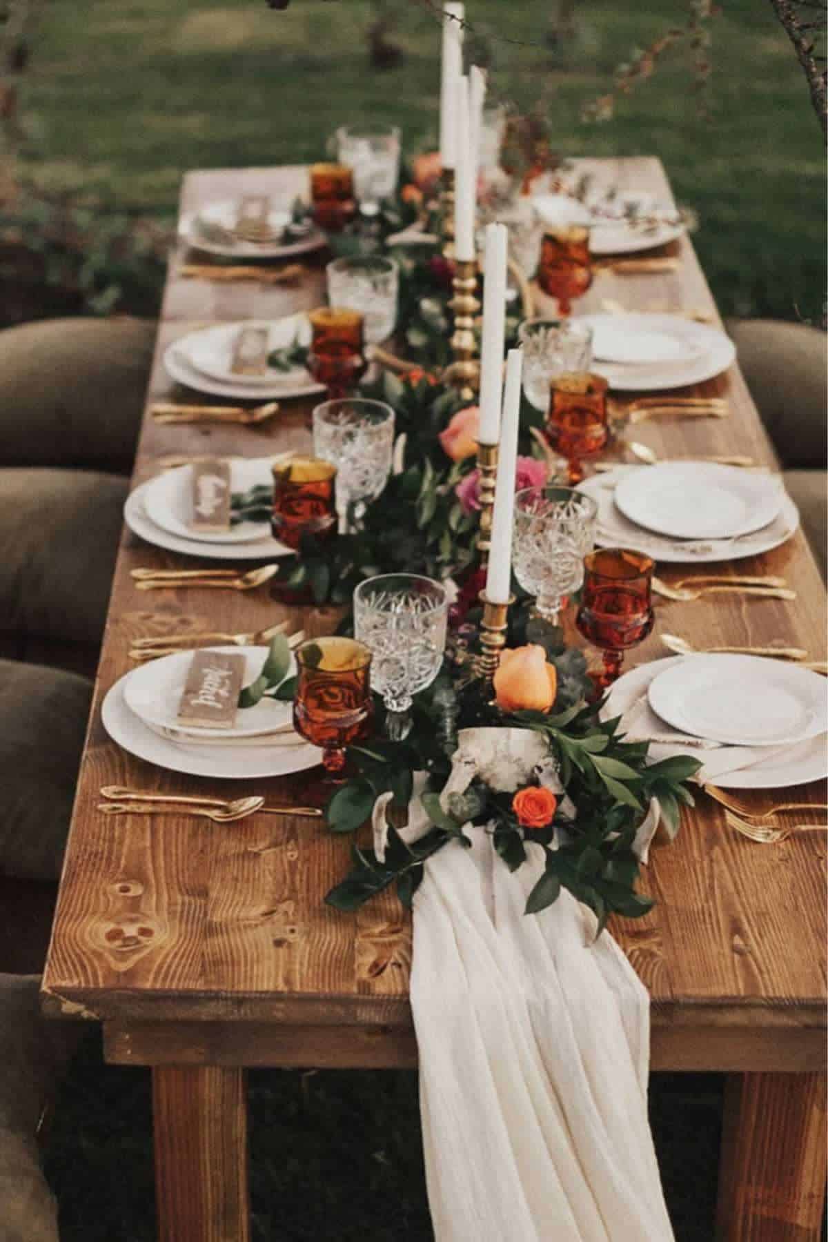 A rustic wooden table is set outdoors with plates, crystal glasses, gold cutlery, candles, floral centerpieces, and a white table runner, ready for a meal.