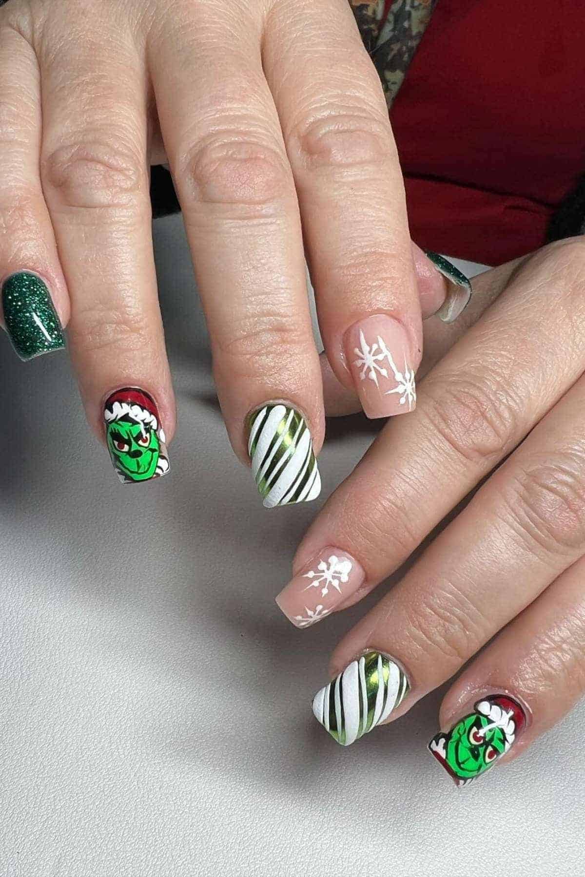 Hands with manicured nails featuring green glitter, white snowflakes, striped patterns, and designs of the Grinch character.