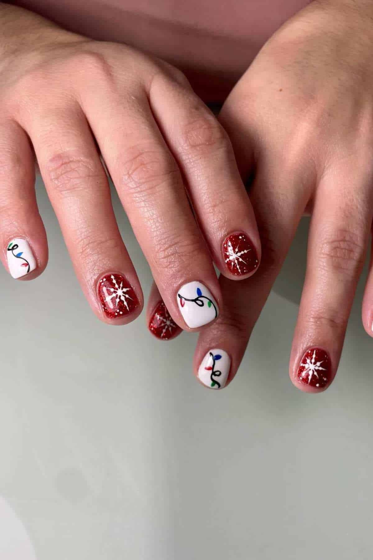Hands with red glitter nail polish, white snowflake designs on some nails, and multicolored Christmas light designs on white accent nails.