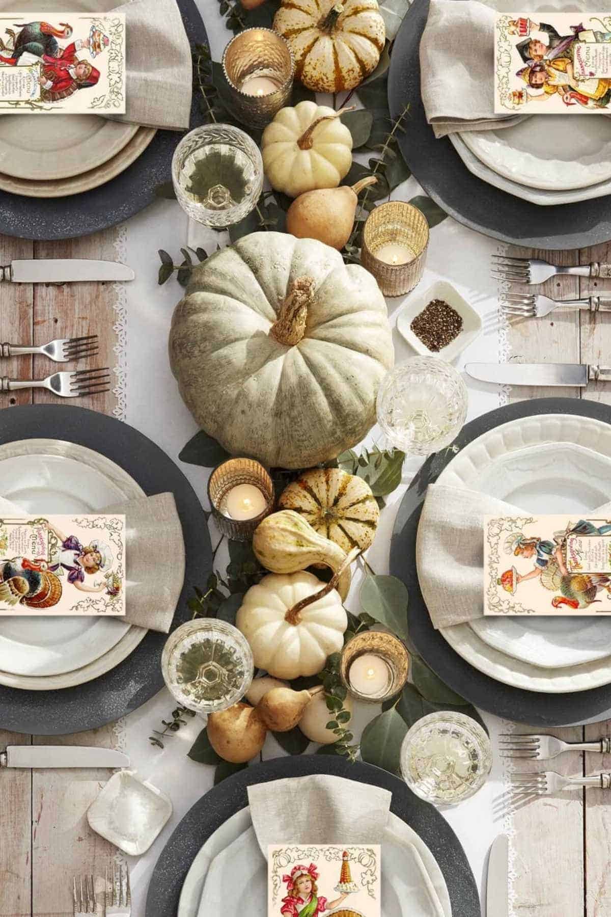 A fall-themed table setting with white and green pumpkins, candles, greenery, plates, cutlery, glasses, and illustrated cards arranged around a rustic centerpiece.
