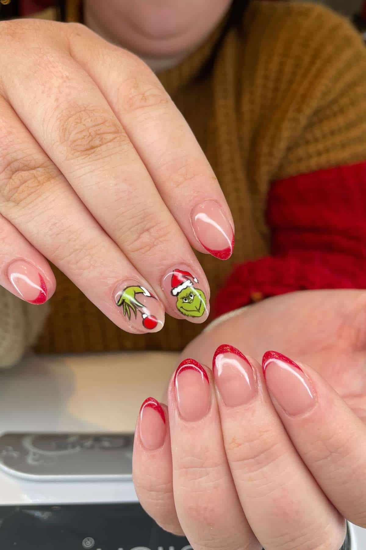 A person displays manicured nails with red French tips; two nails feature holiday-themed art, including a Santa hat, the Grinch, and a green hand holding an ornament.