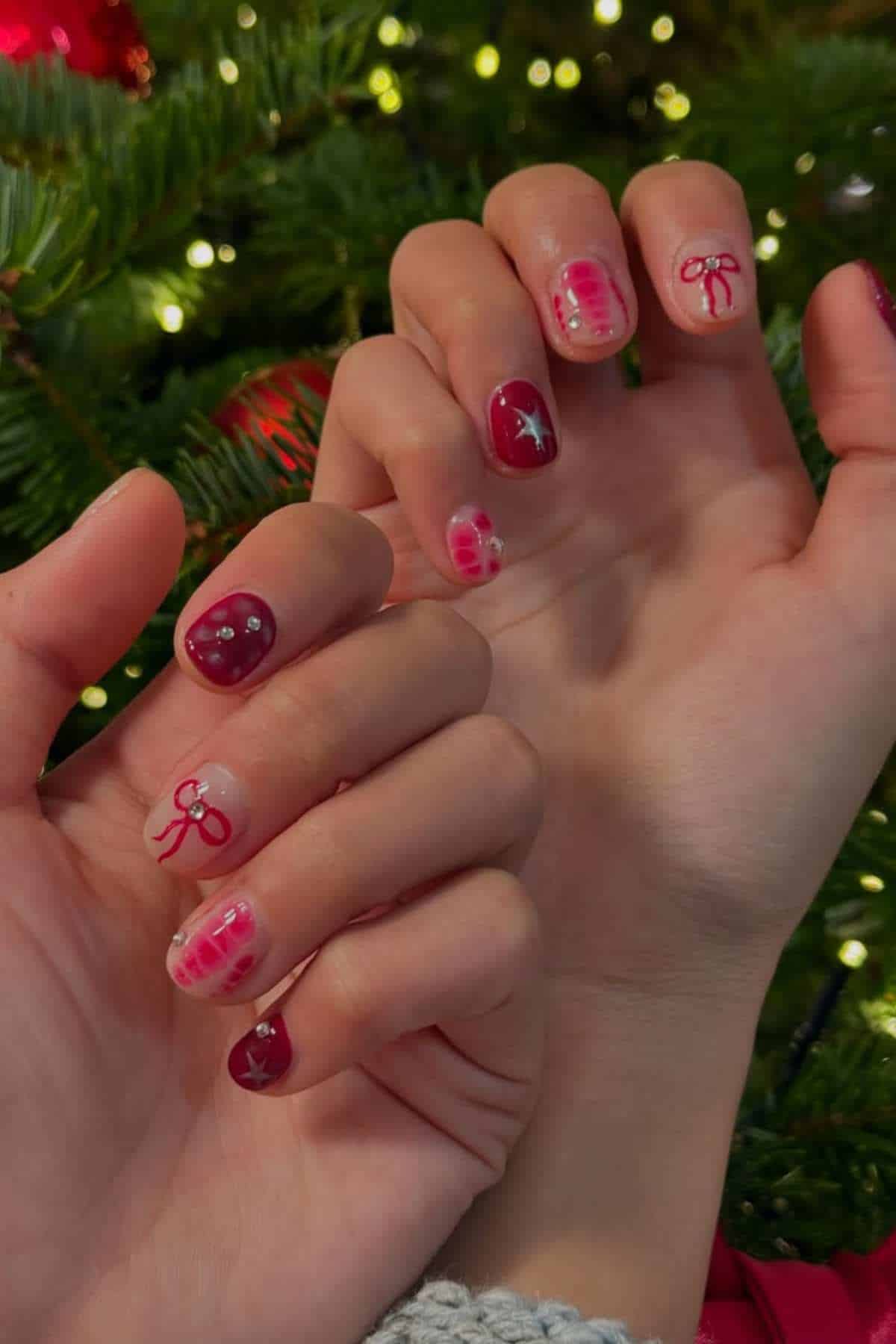Close-up of hands with festive nail art featuring red polish, bows, and star designs in front of a decorated Christmas tree.