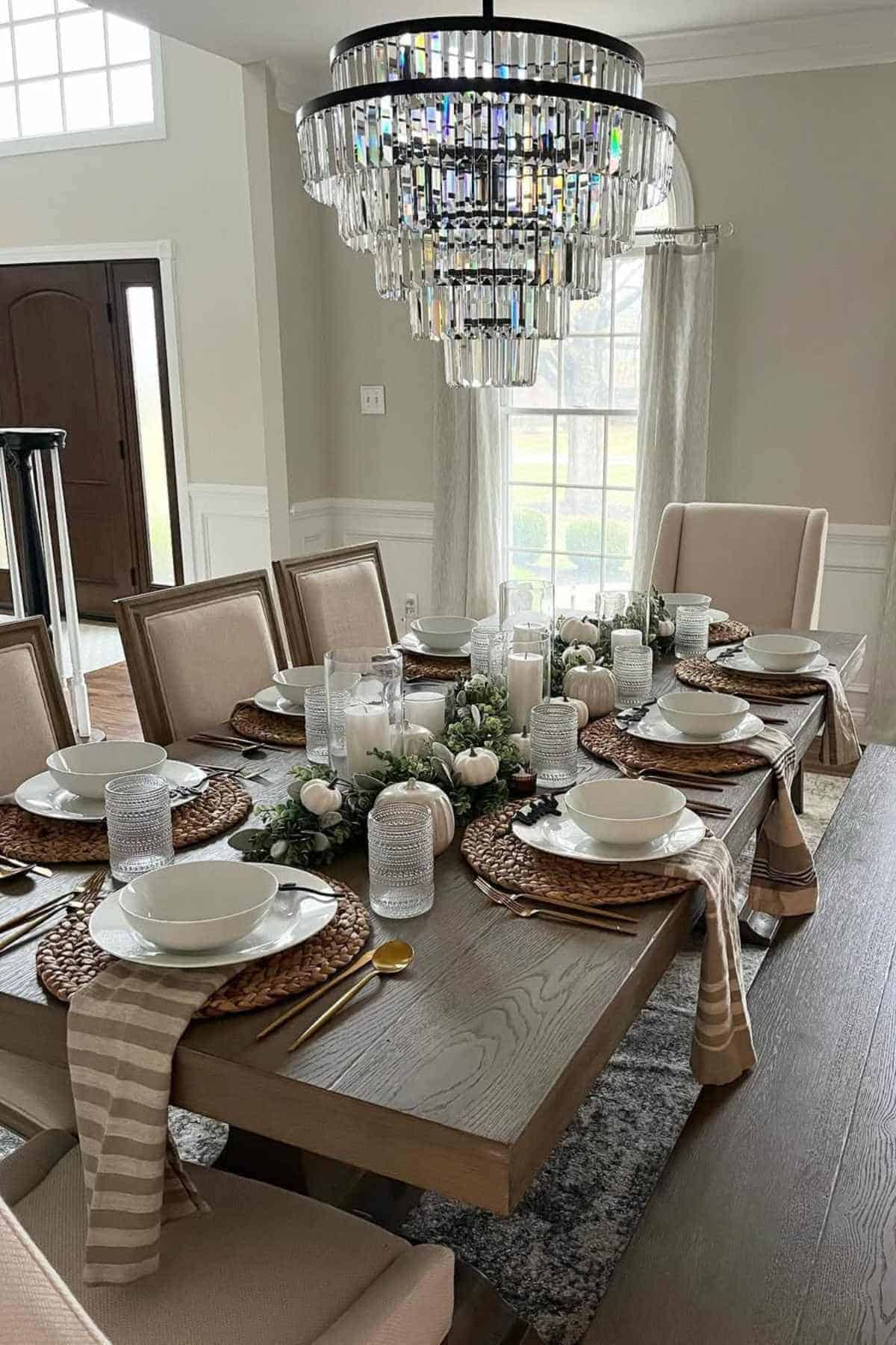 A dining room with a wooden table set for six, featuring neutral tableware, woven placemats, striped napkins, candles, and a modern crystal chandelier overhead.