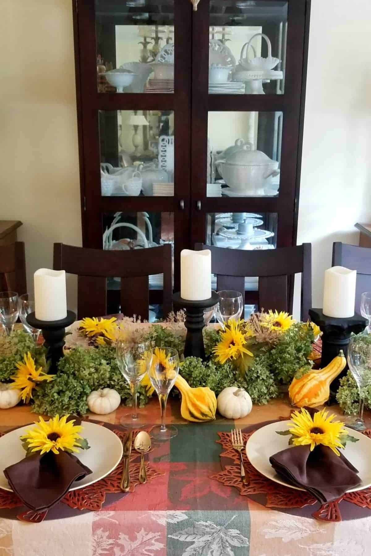 A dining table set with white plates, brown napkins, sunflowers, candles, gourds, and greenery, with a cabinet of white dishware in the background.