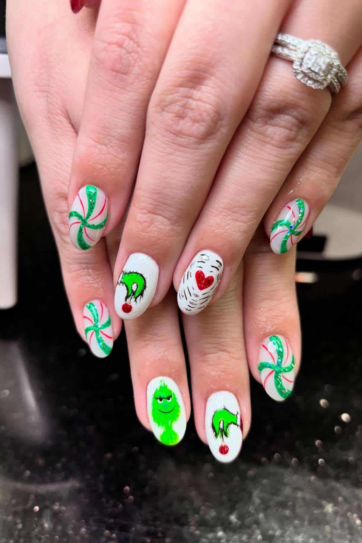 A hand with white manicured nails featuring green and red holiday designs, including peppermint swirls, the Grinch, and a heart.