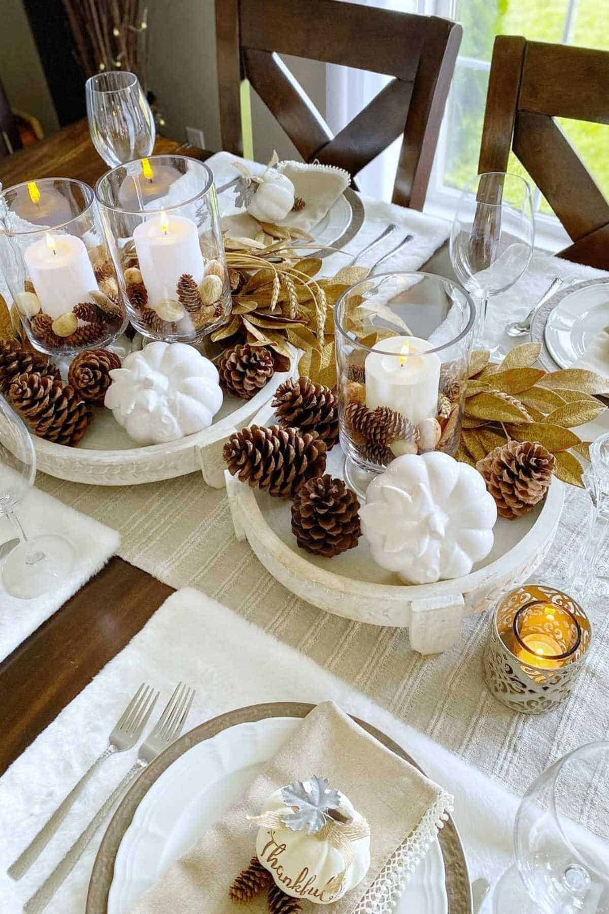 A dining table set with white plates, pinecones, white pumpkins, gold leaves, candles in glass holders, and napkins labeled "thankful" in a fall-themed centerpiece arrangement.