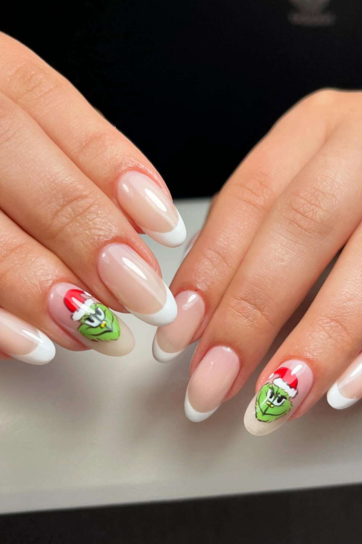 Hands with a French manicure; one nail on each hand features an accent nail art of the Grinch wearing a red Santa hat.