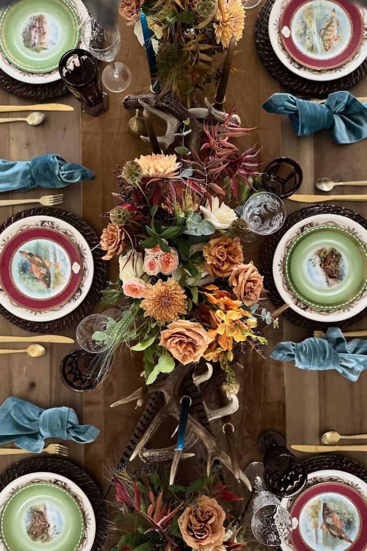 A wooden dining table set with green and cream plates, gold cutlery, blue napkins, and a centerpiece of assorted autumn flowers and antlers.