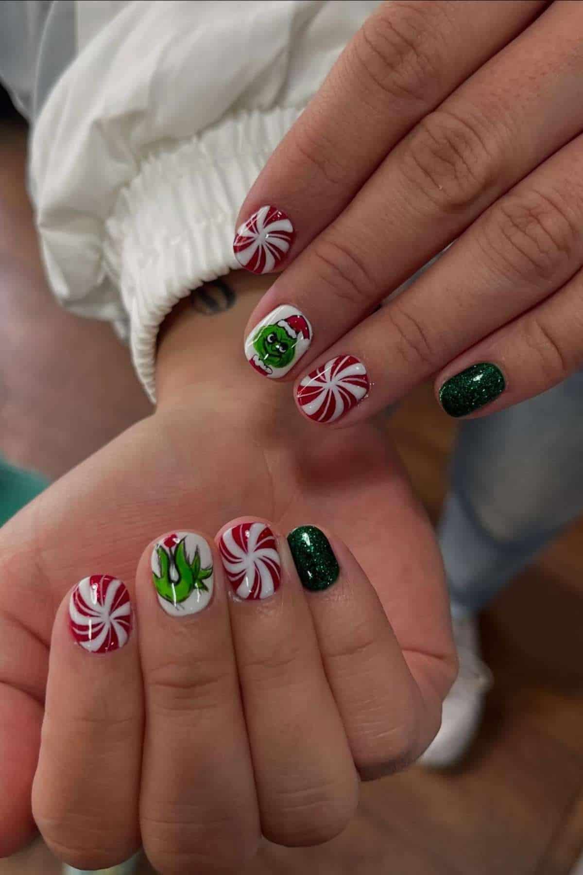 A close-up of hands with festive nail art featuring green glitter, red and white peppermint swirls, and the Grinch character wearing a Santa hat.