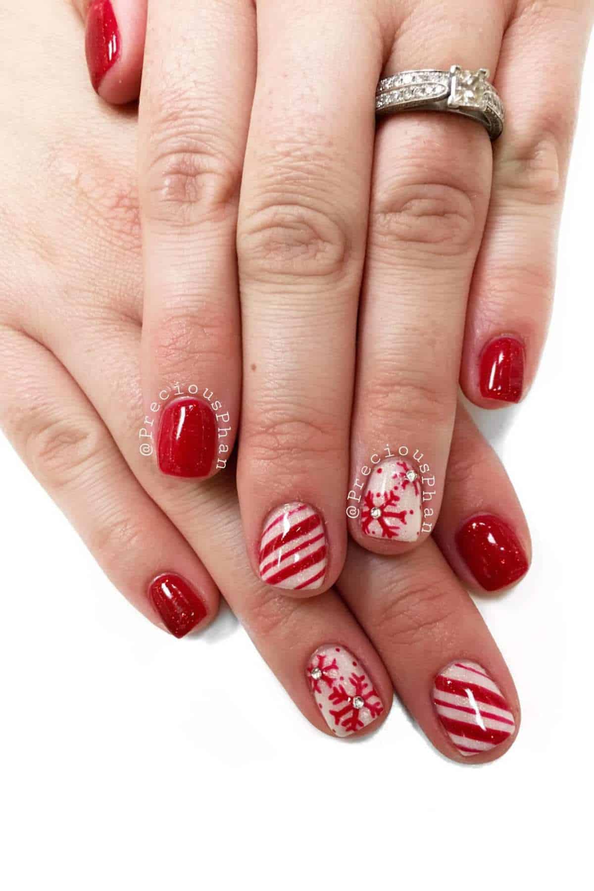Hands with glossy red nail polish; ring fingers feature white and red snowflake and striped designs. Person is wearing a silver ring with a square-cut gemstone.
