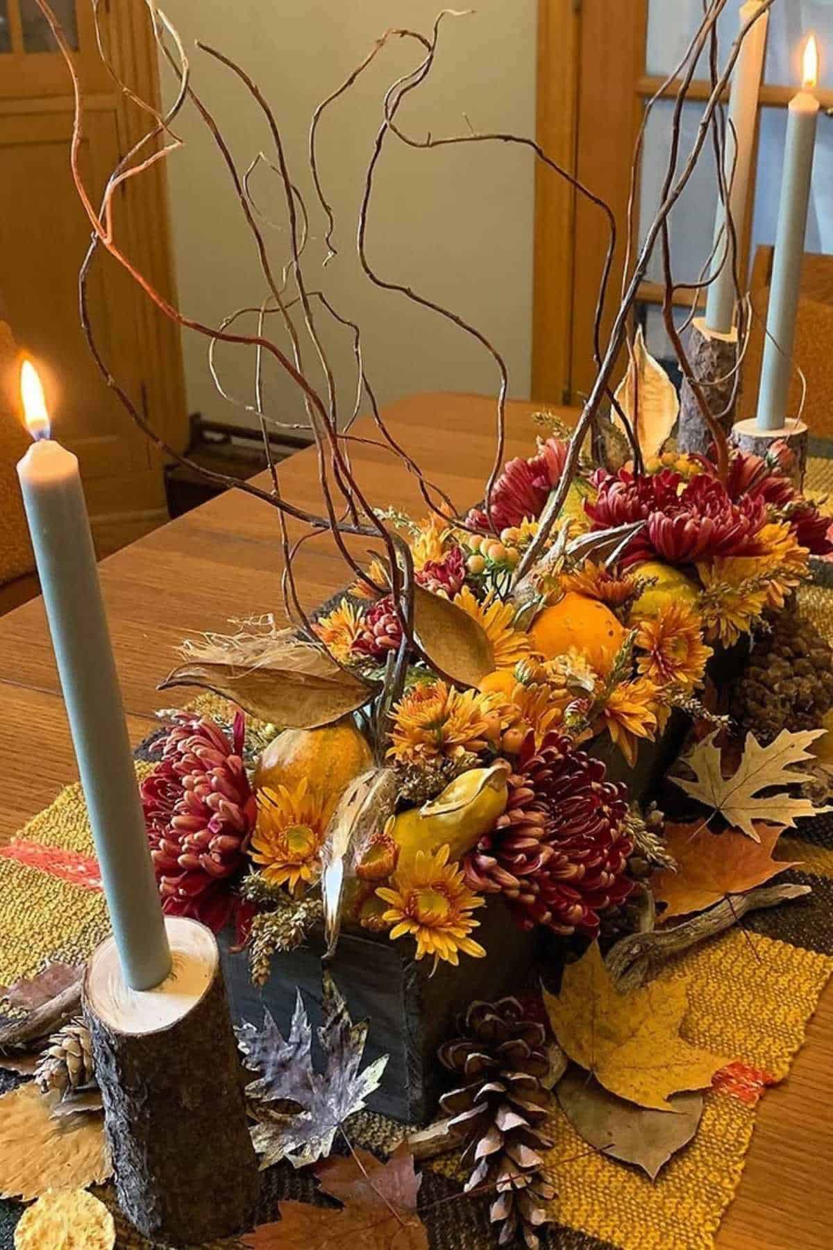 A fall-themed table centerpiece with pumpkins, colorful flowers, pinecones, autumn leaves, and tall branches, flanked by two lit candles in wooden holders.
