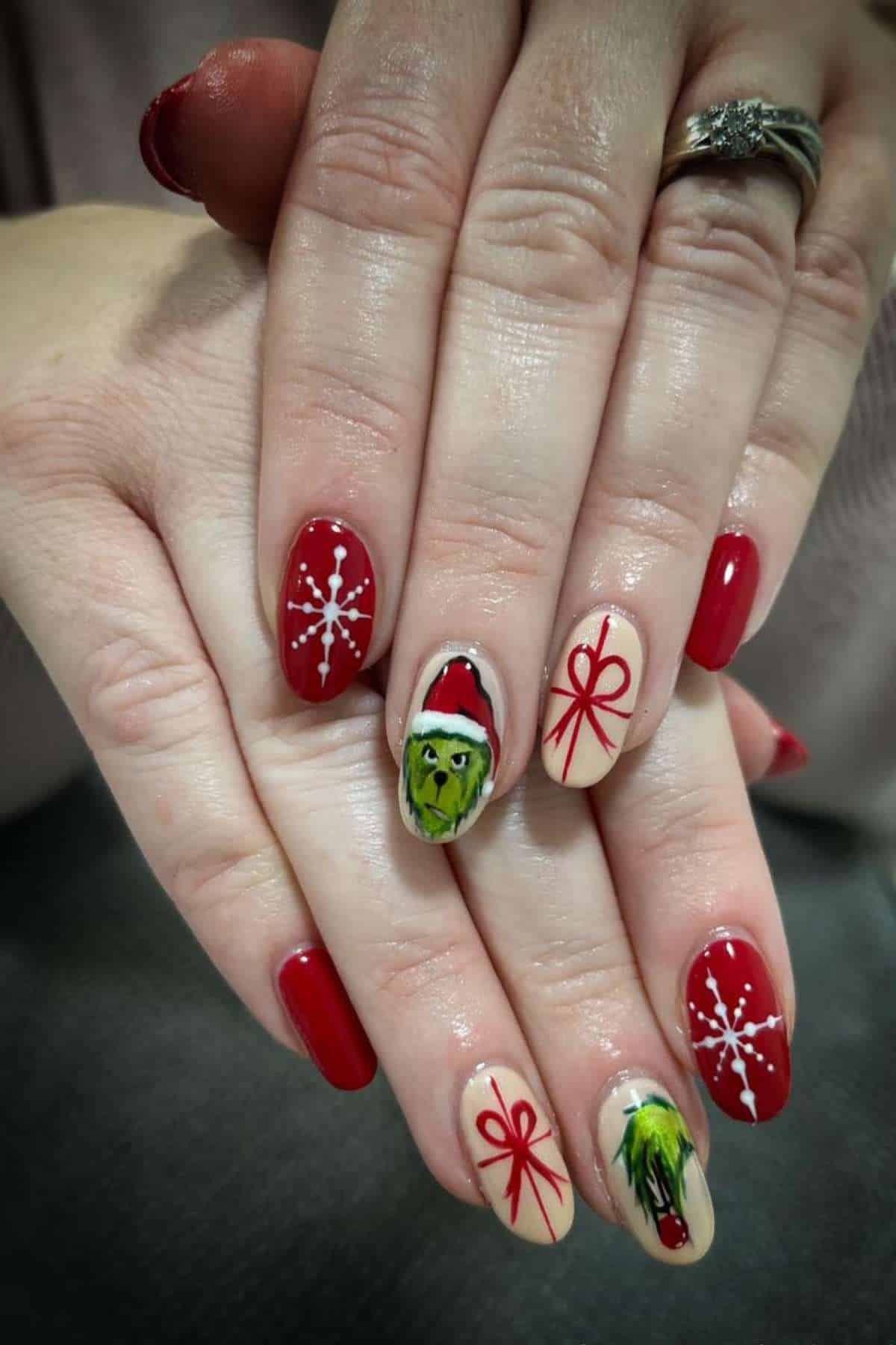 Hands with red and nude Christmas-themed nail art, featuring snowflakes, gift bows, and the Grinch character wearing a Santa hat.