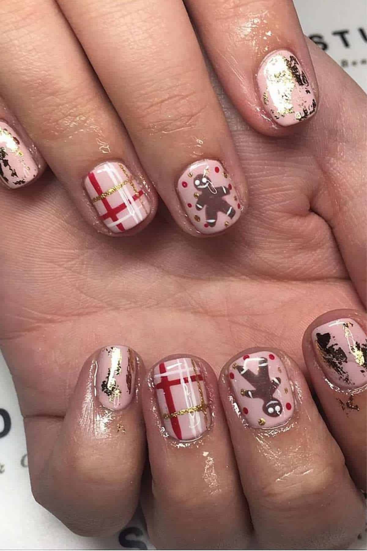 Close-up of hands with nail art featuring plaid patterns, gold accents, and gingerbread man designs on a light pink base.