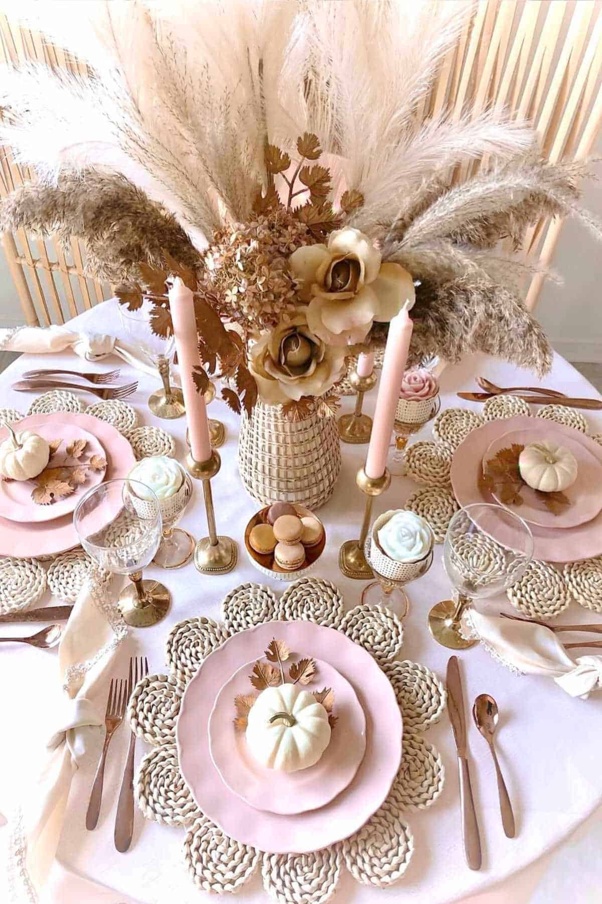 A table set for four with pink plates, gold cutlery, small white pumpkins, woven placemats, candles, and a centerpiece of dried flowers and pampas grass.
