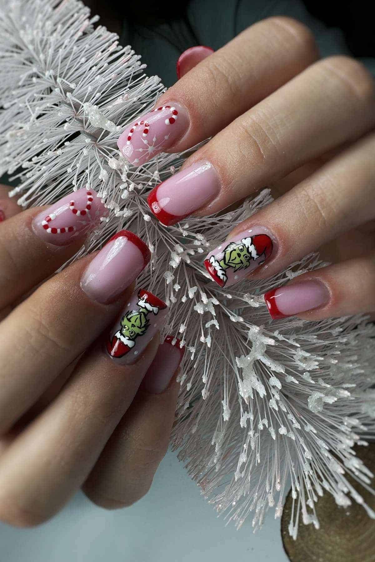 A close-up of hands with festive nail art featuring red tips, heart designs, and The Grinch wearing a Santa hat, holding a white decorative branch.