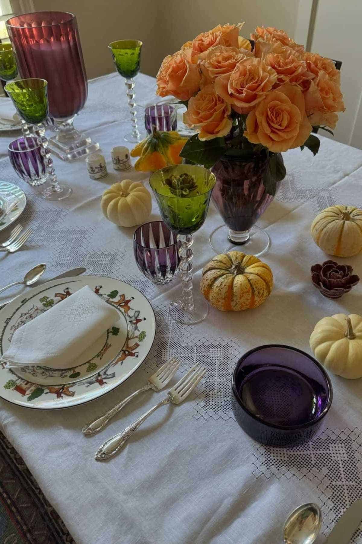 A table set with plates, silverware, colored glassware, a vase of orange roses, and small decorative pumpkins on a white lace tablecloth.