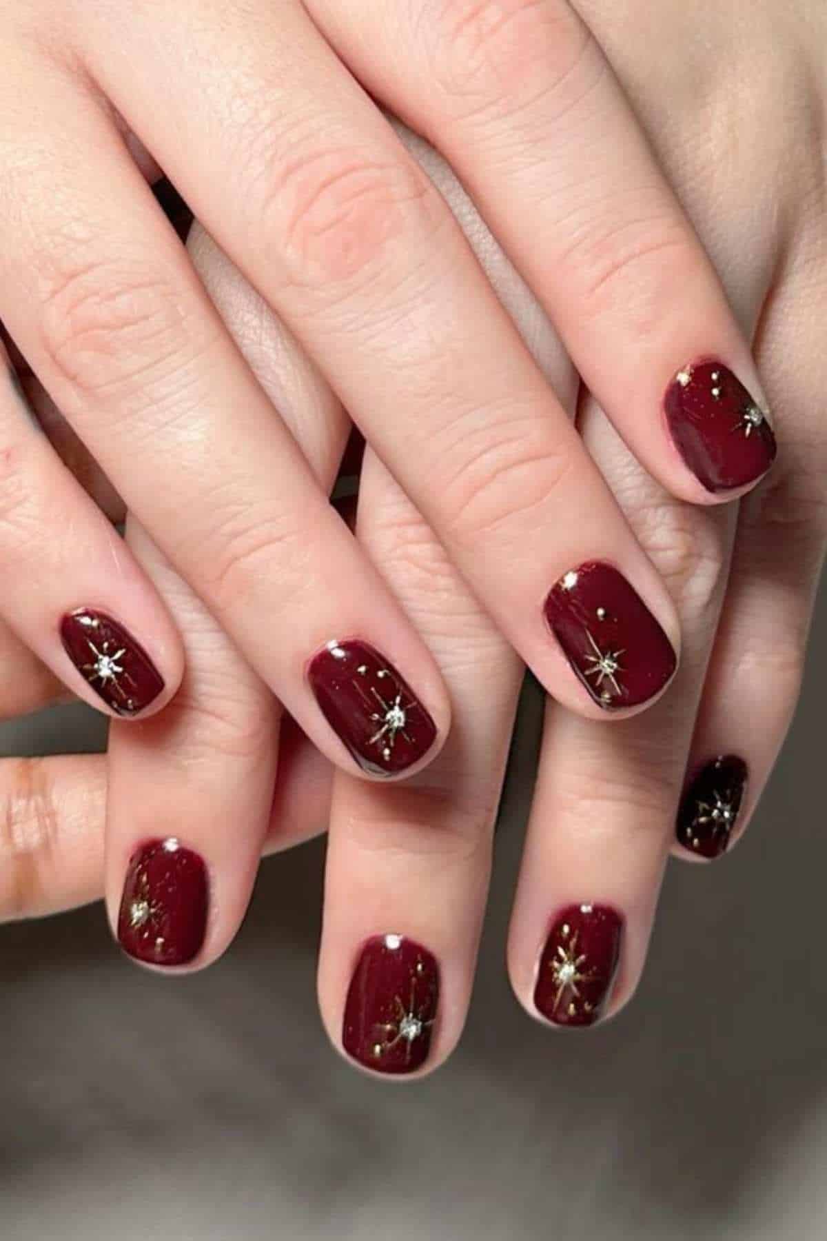 Hands with short, oval-shaped nails painted dark red, each nail decorated with a small gold starburst design.