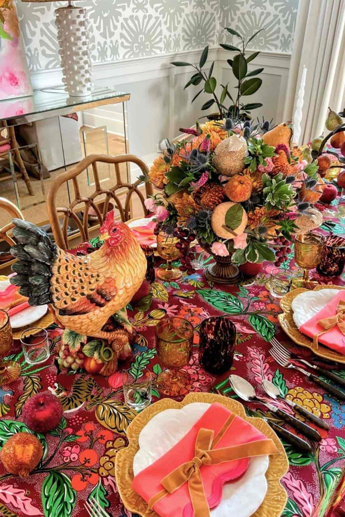 A dining table is set with a vibrant floral tablecloth, a ceramic hen centerpiece, floral arrangement, plates, gold cutlery, and orange napkins in a bright, decorated room.
