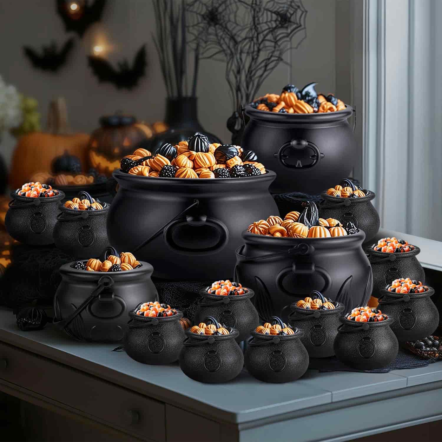 A group of black cauldrons filled with assorted Halloween candies sits on a table, with Halloween decorations and pumpkins in the background.