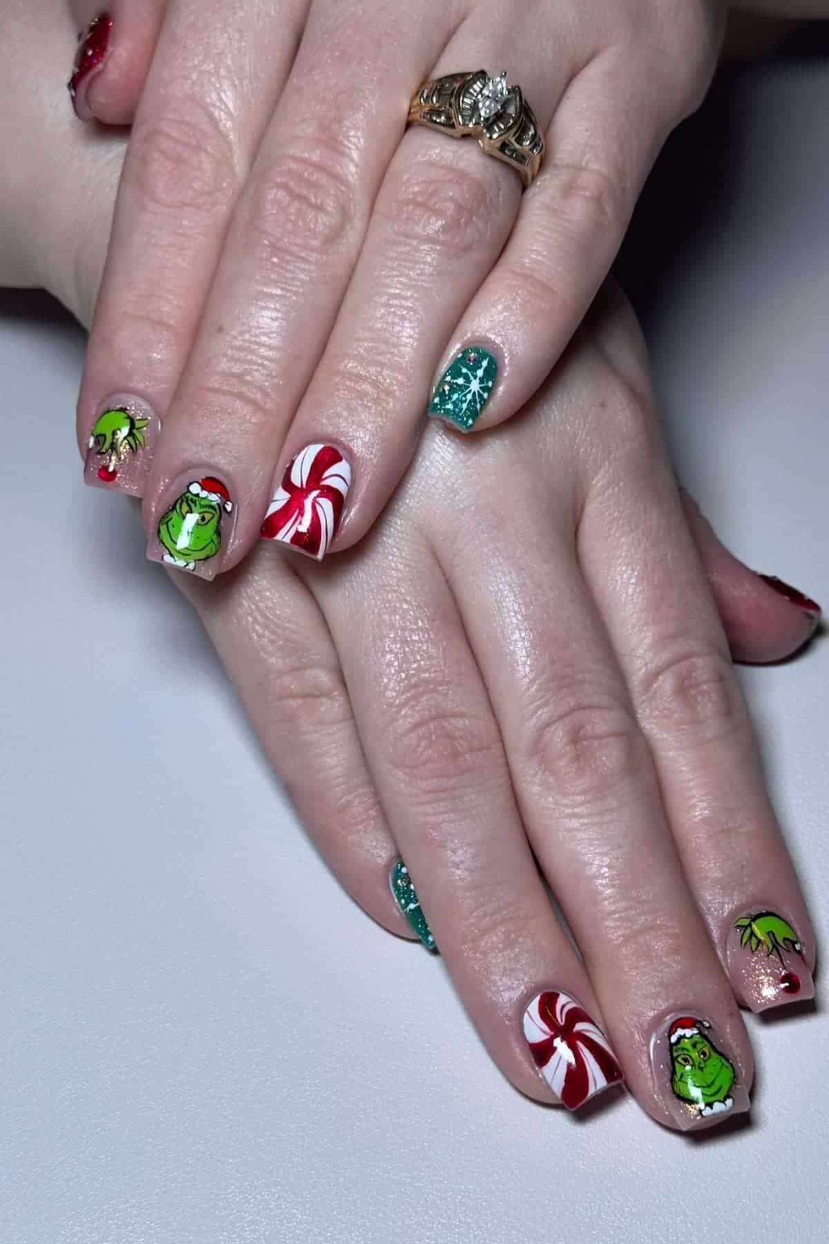 Hands with festive nail art featuring the Grinch, candy canes, holly leaves, and sparkling green polish. The person is wearing a gold ring with clear stones.