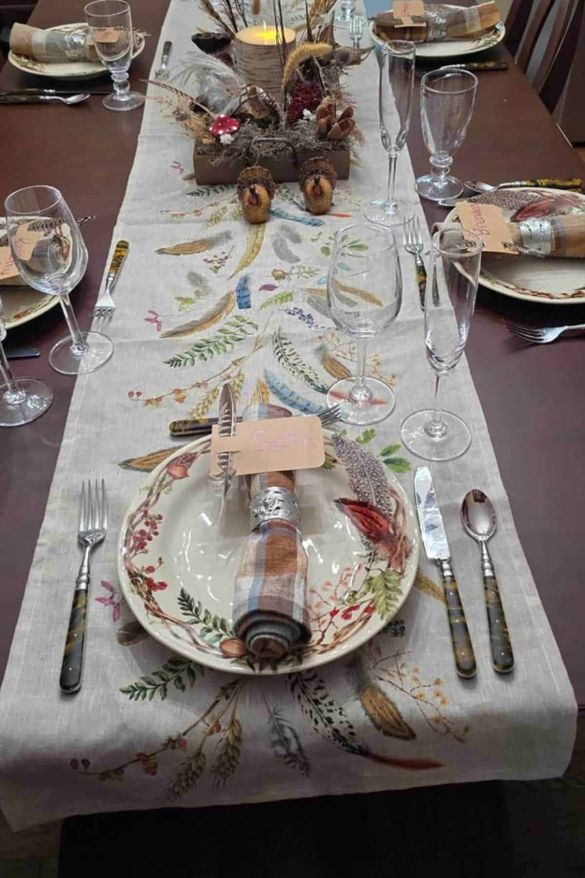 A dining table set for a meal with plates, glasses, silverware, plaid napkins, and a nature-themed table runner featuring leaves and feathers. A rustic centerpiece is displayed.