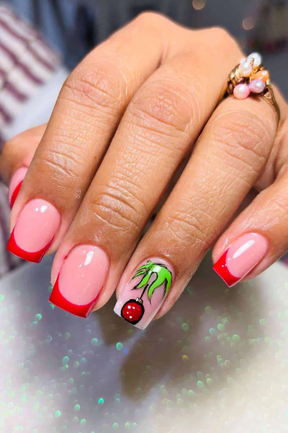 A hand with pink and red French tip nails; the ring fingernail features the hand of the grinch and red glitter design.