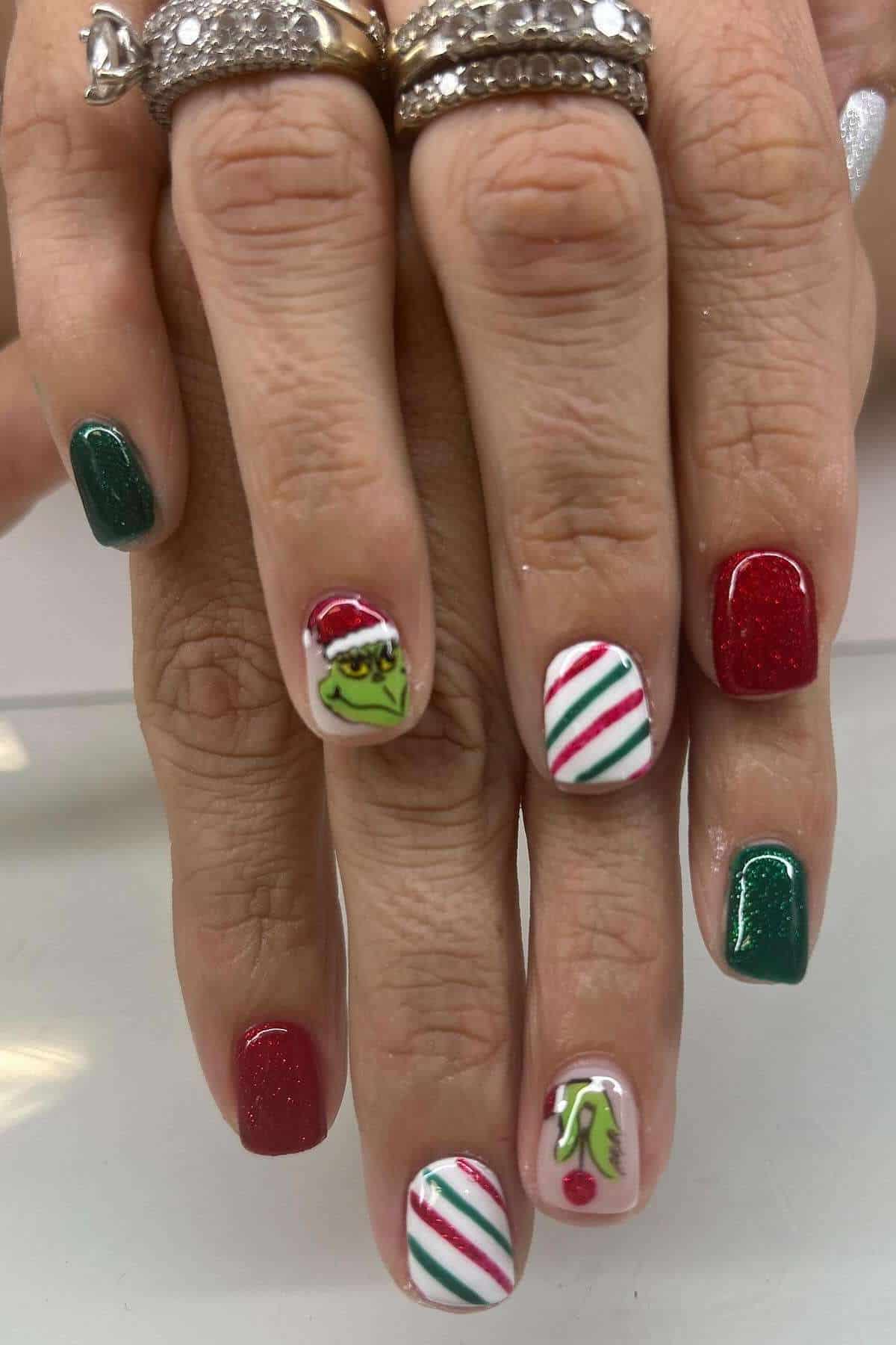 Two hands with festive nail art: alternating red and green glitter polish, candy cane stripes, and designs of the Grinch's face and hand wearing a Santa hat.