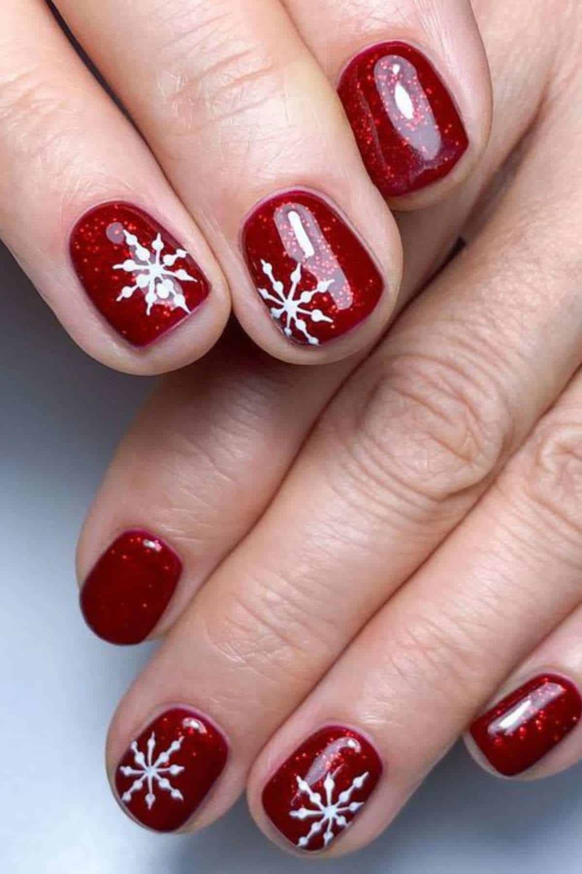 Hands with short, red painted nails featuring white snowflake designs on some nails.