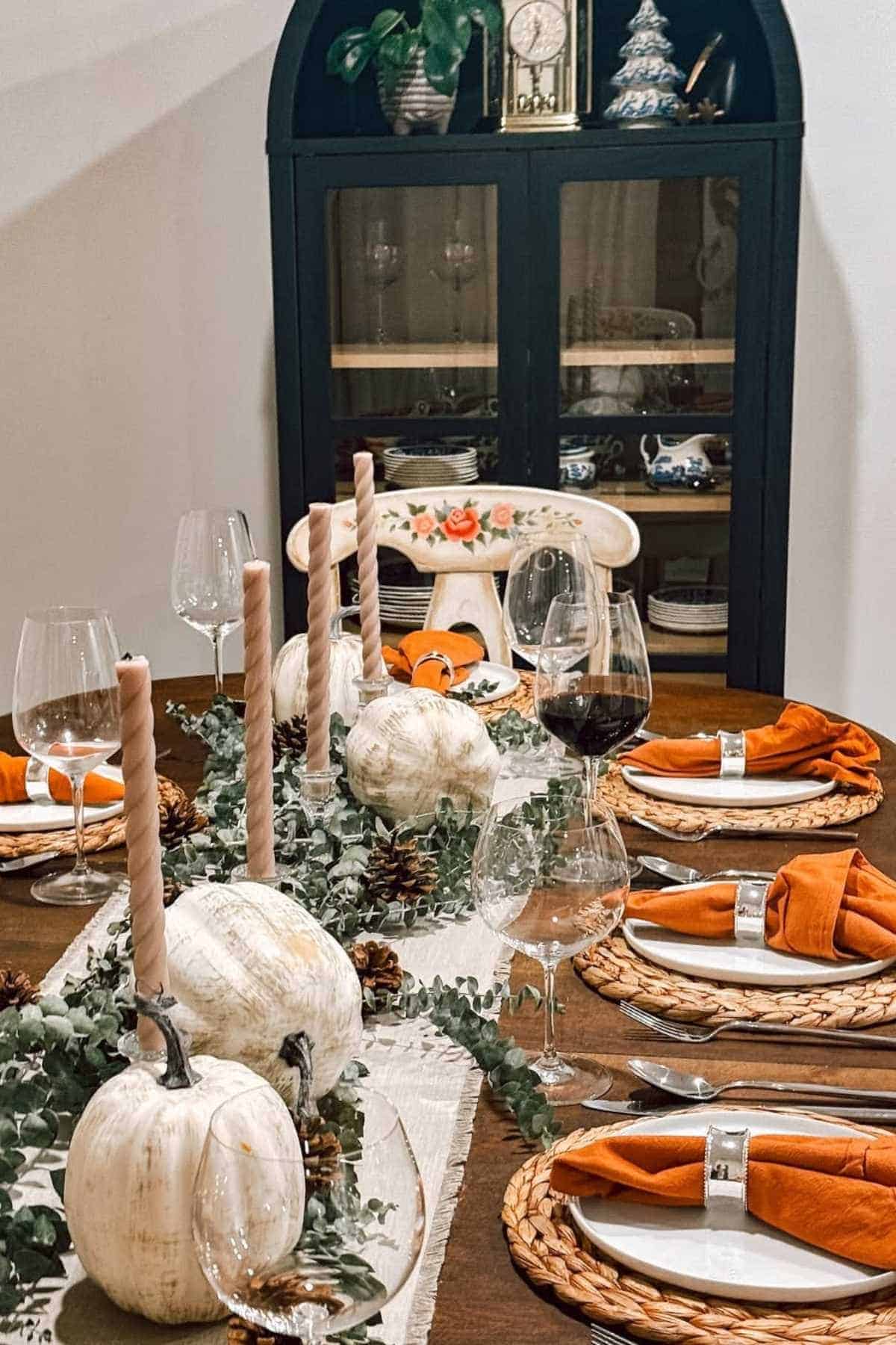 A dining table set for a meal features white pumpkins, tapered candles, orange napkins, and glassware, with a cabinet and decorative items in the background.