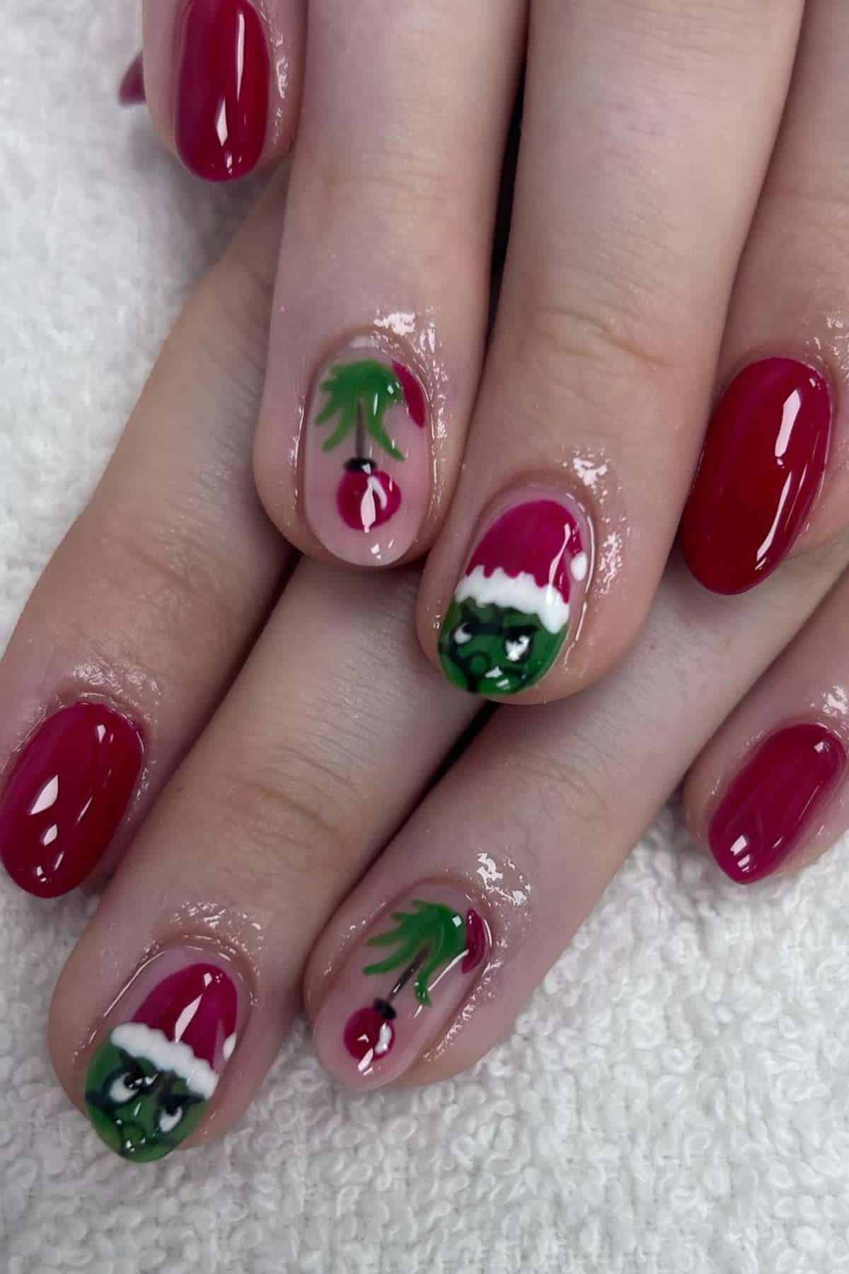 Hands with red nail polish; two nails feature festive designs: one with the Grinch in a Santa hat, and the other with a cherry and green leaf.