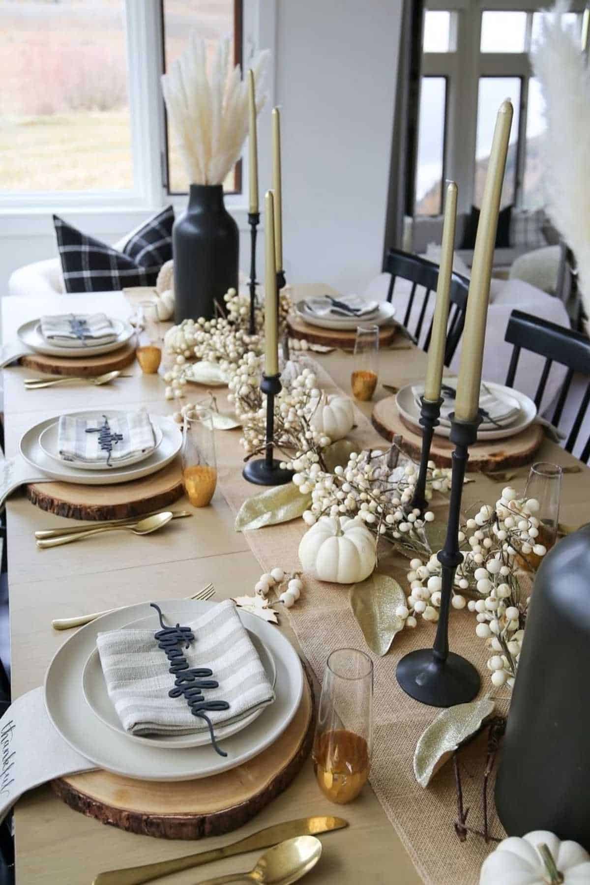 A dining table set for a meal with neutral-toned plates, black candlesticks, white pumpkins, dried floral decorations, and glasses on a wooden table.