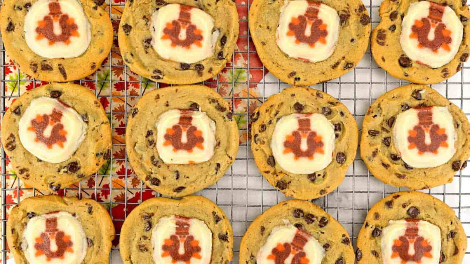 A cooling rack holds twelve chocolate chip cookies, each topped with a round slice of marshmallow decorated with a red maple leaf design.
