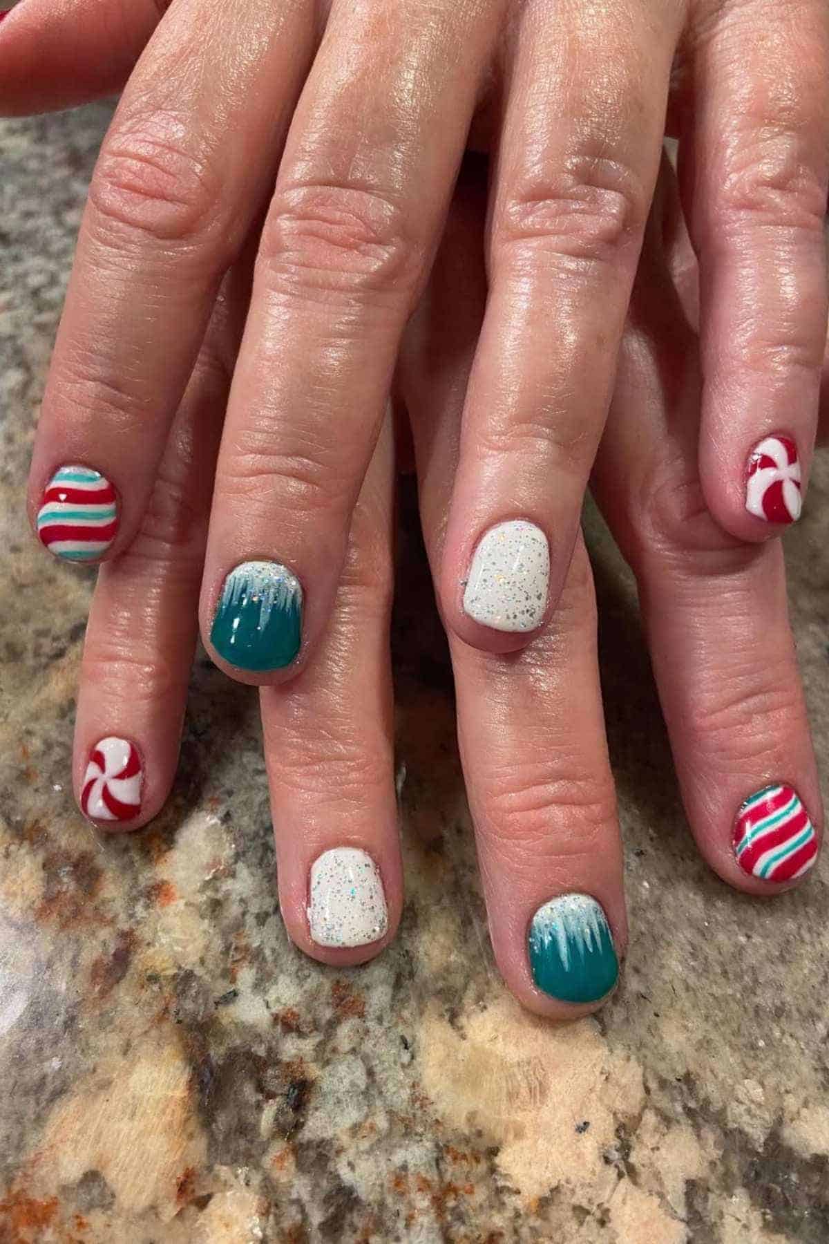 A hand with festive nail art featuring red and white stripes, turquoise tips, glittery white, and peppermint swirl designs, displayed on a granite countertop.