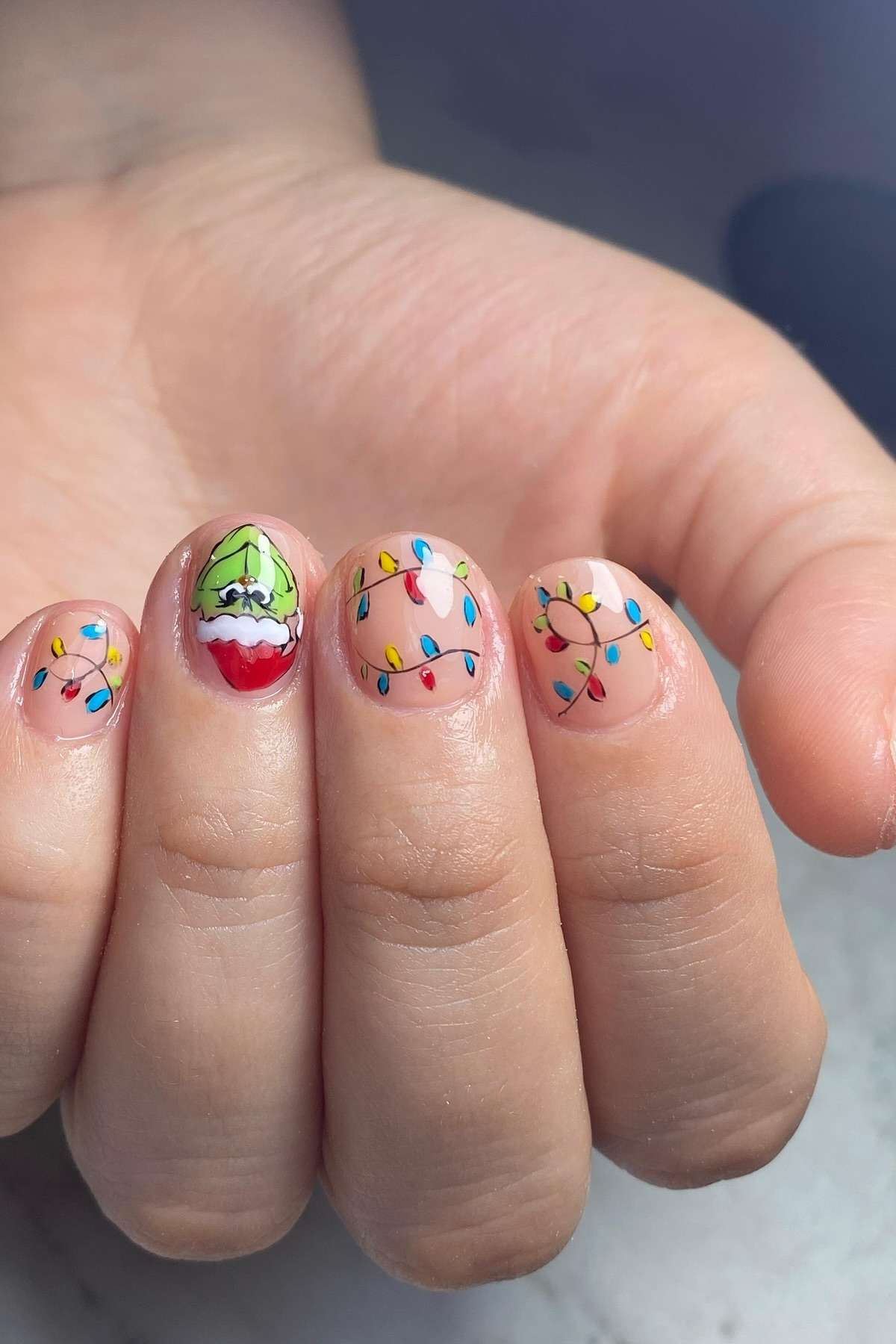 A hand with festive nail art featuring colorful Christmas lights on most nails and the Grinch's face in a Santa hat on the ring finger.