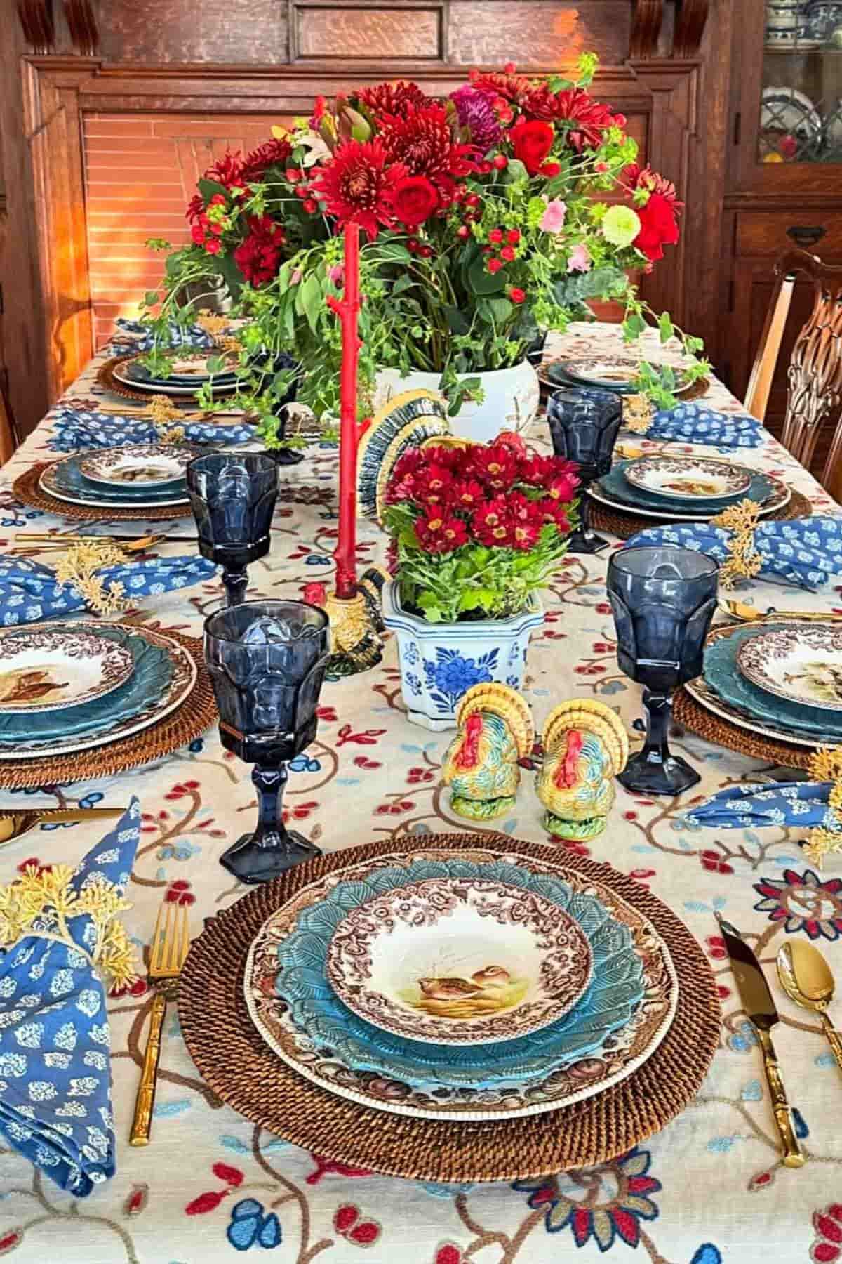 A dining table set with patterned plates, blue glasses, floral centerpiece, turkey figurines, gold cutlery, and a tablecloth with a floral design.