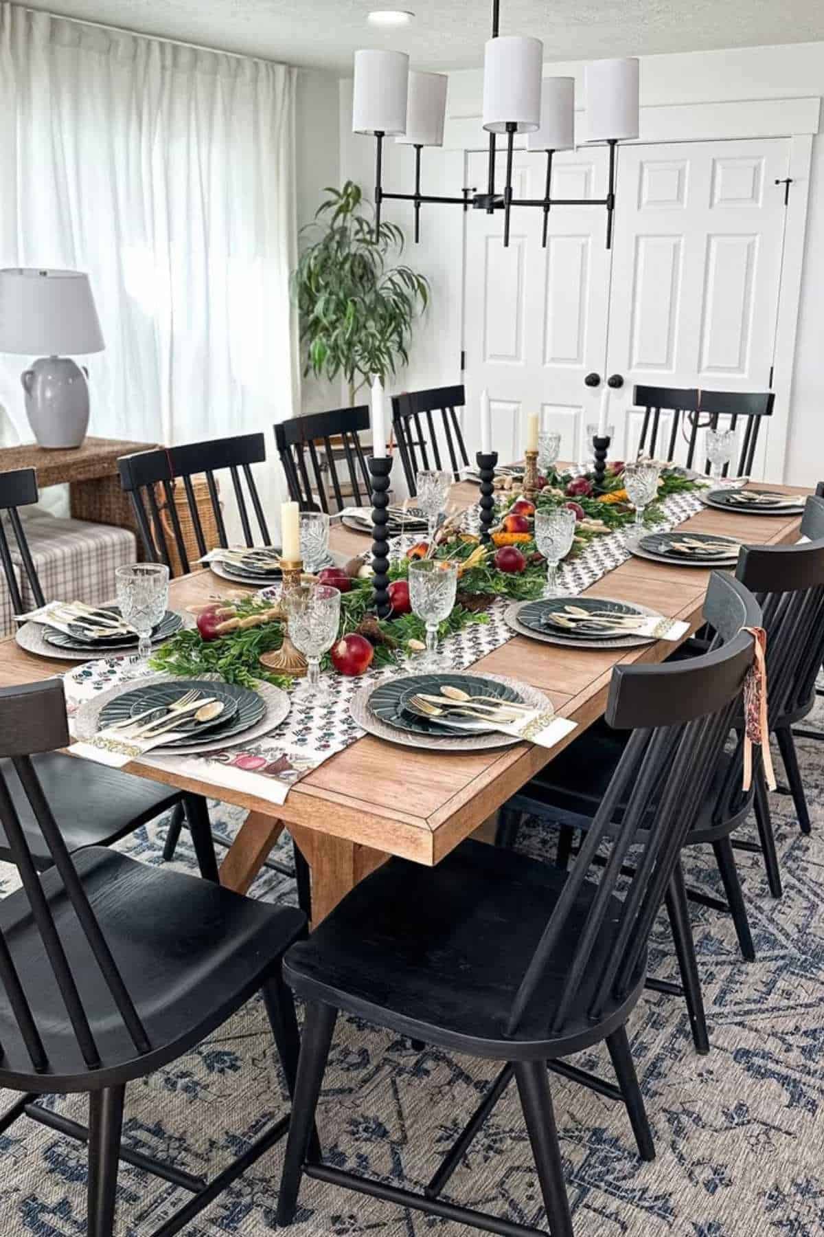 A dining table set for ten with black chairs, candles, plates, glasses, cutlery, greenery, apples, and a patterned table runner under a modern chandelier.