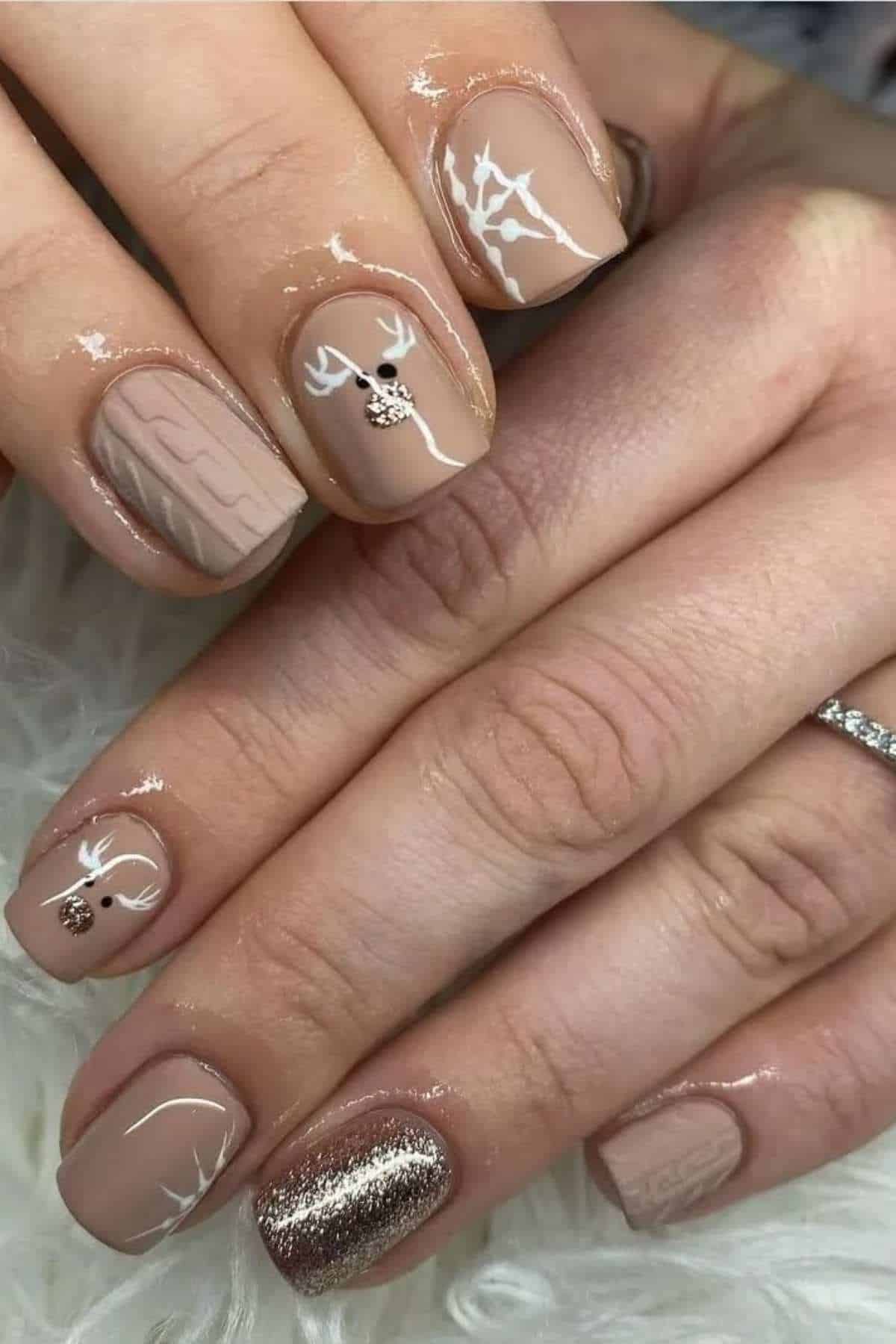 A hand with nude-colored nails featuring white reindeer designs, a knit pattern, a gold glitter accent nail, and a silver ring on the ring finger.