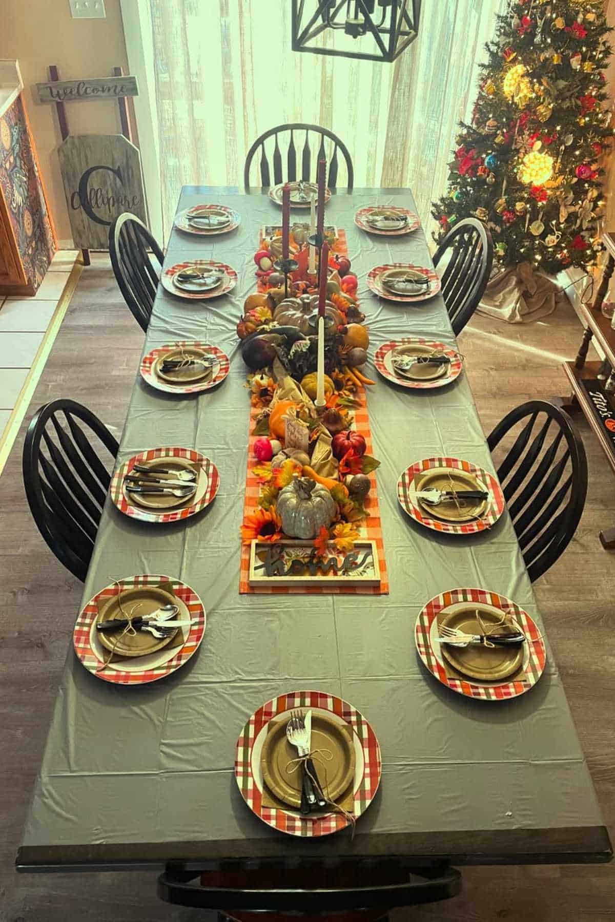 A dining table set for twelve with plaid-rimmed plates, silverware, and a fall-themed centerpiece; a decorated Christmas tree is visible in the background.