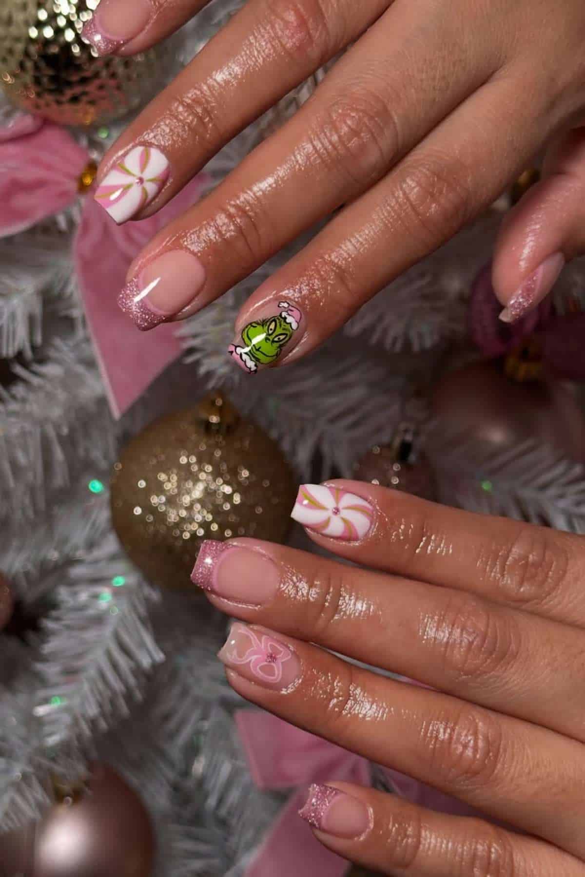 Close-up of hands with manicured nails, featuring pink French tips, pinwheel designs, and one accent nail with a frog face. A white Christmas tree and ornaments are in the background.