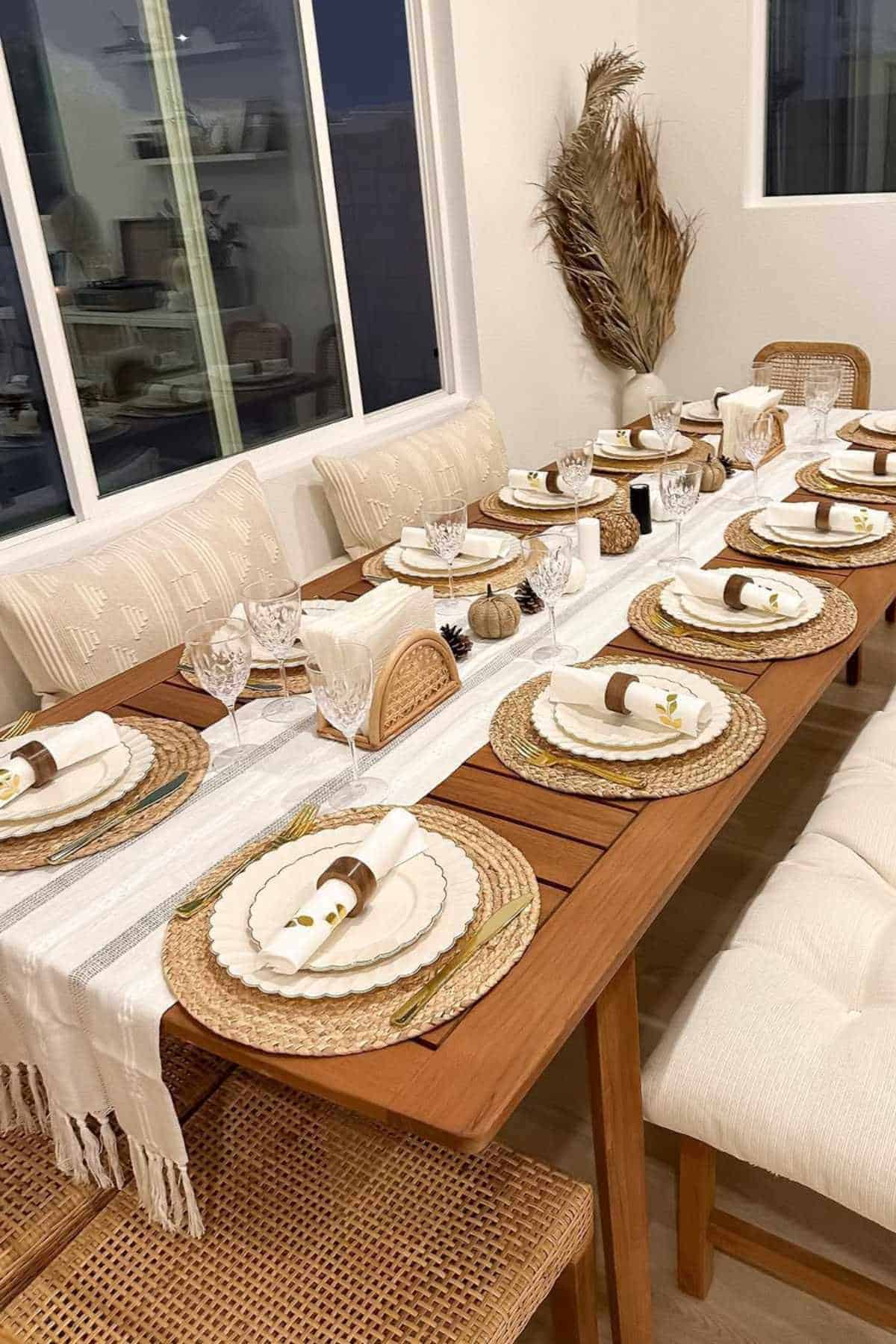 A wooden dining table set for eight with woven placemats, white plates, rolled napkins, glassware, and neutral-toned decor, next to a window and cushioned seating.