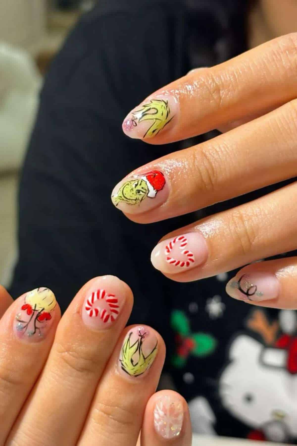 Close-up of hands with festive nail art featuring the Grinch in a Santa hat, peppermint candies, holly, and leaf designs on a light background.