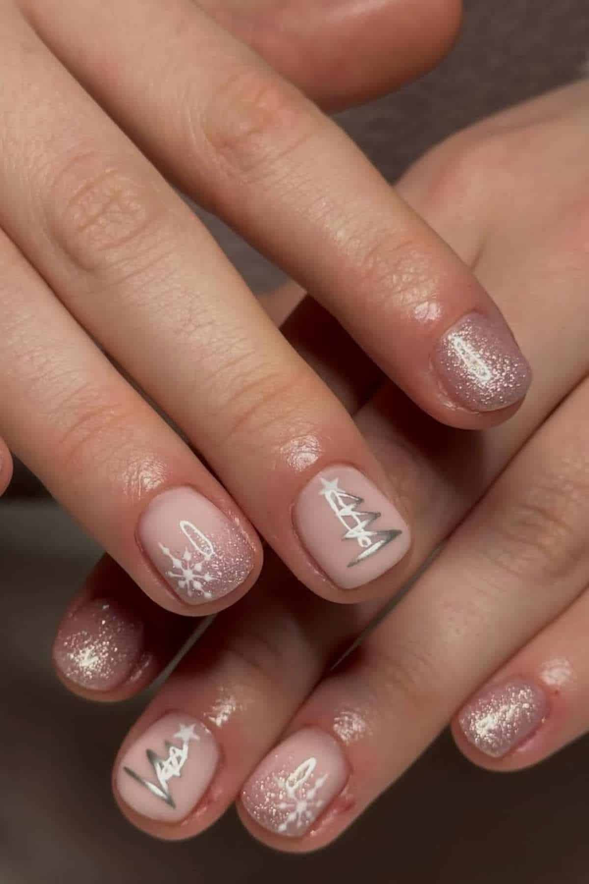 Close-up of hands with short nails featuring glittery pink polish and white winter-themed designs, including snowflakes and tree shapes.