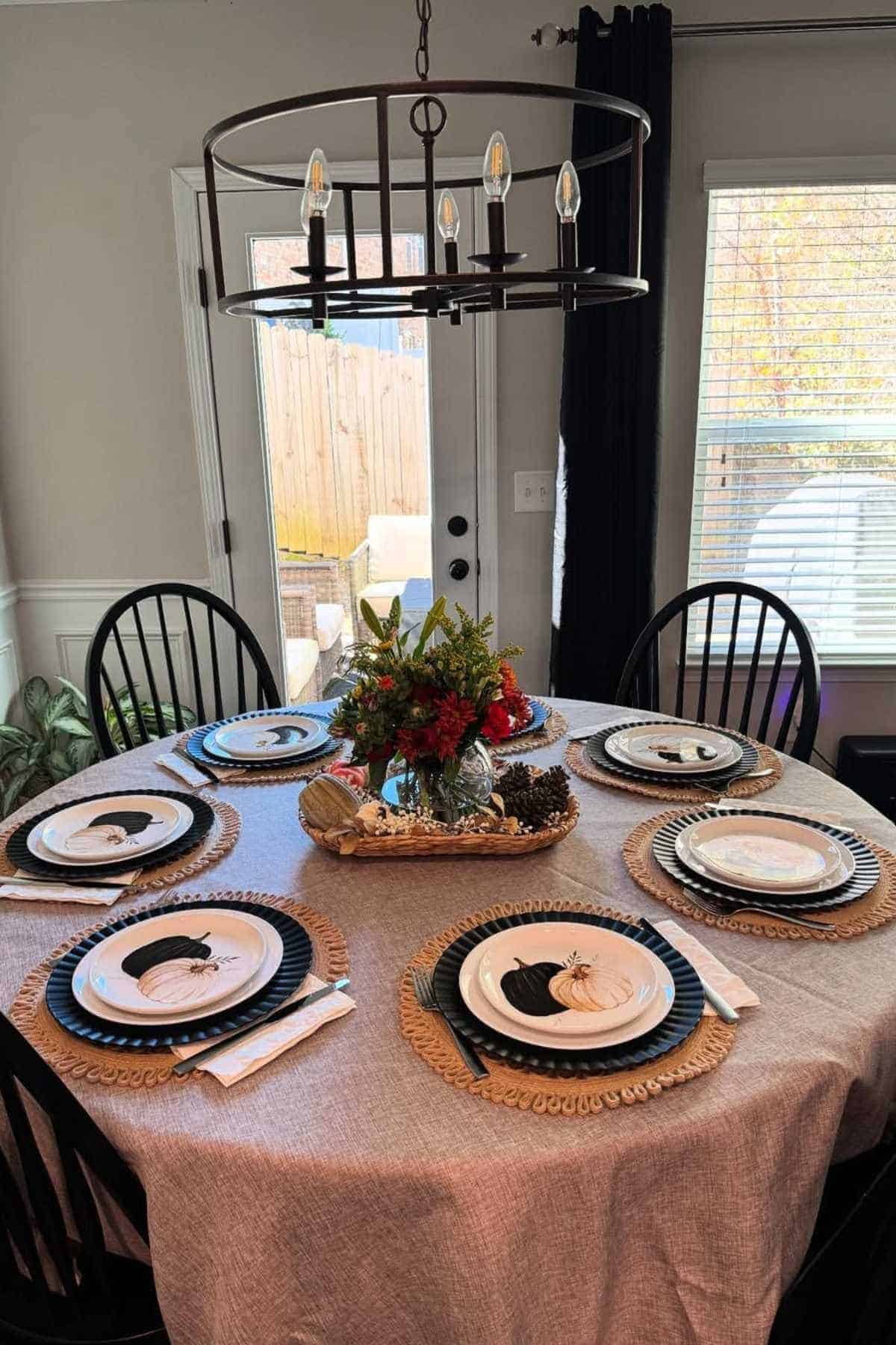 A round dining table set for six with black chairs, autumn-themed plates, a beige tablecloth, and a floral centerpiece under a black chandelier.