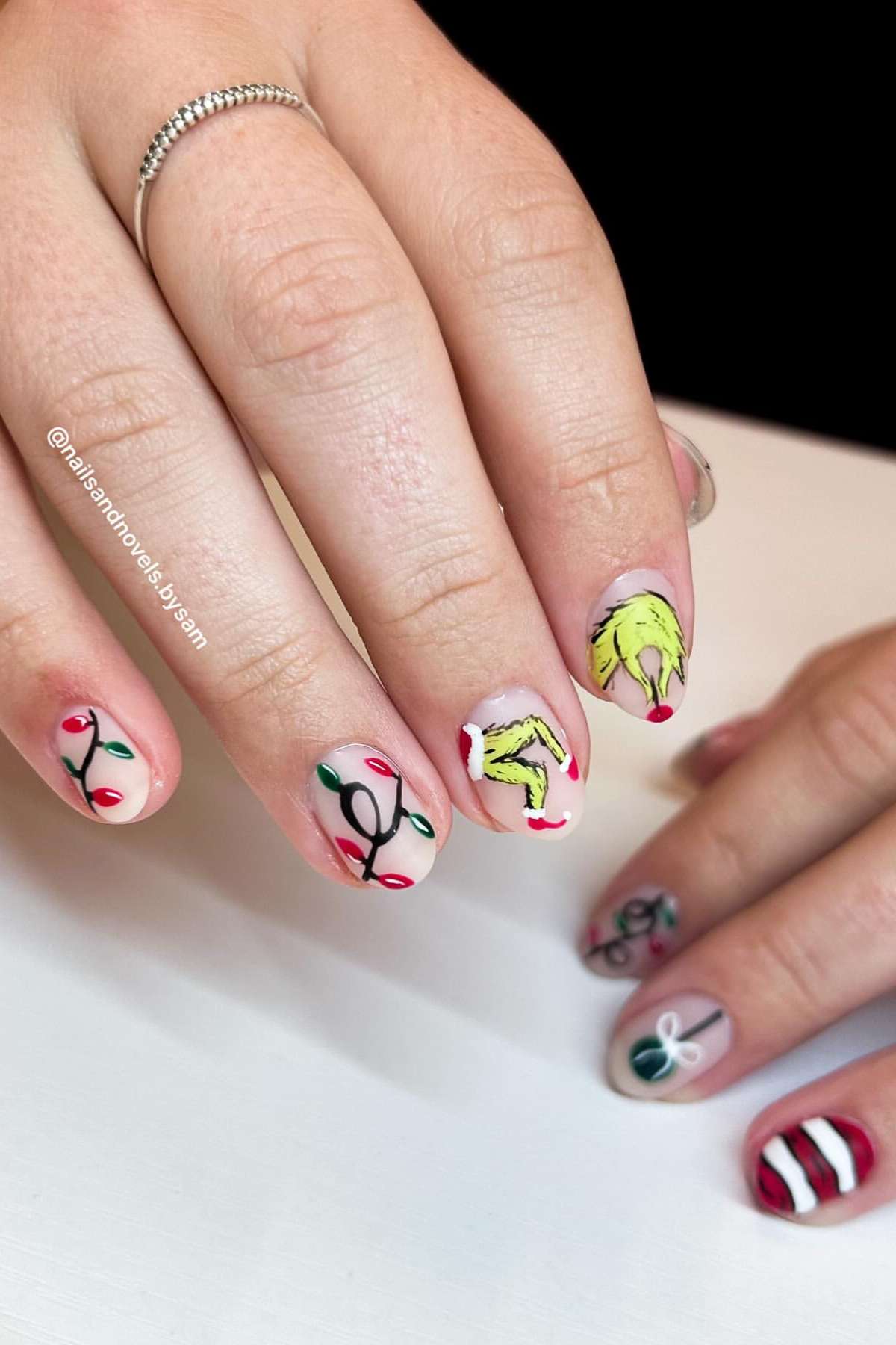 A hand with Christmas-themed nail art, including red and green holiday lights and a design of the Grinch's hand holding an ornament.