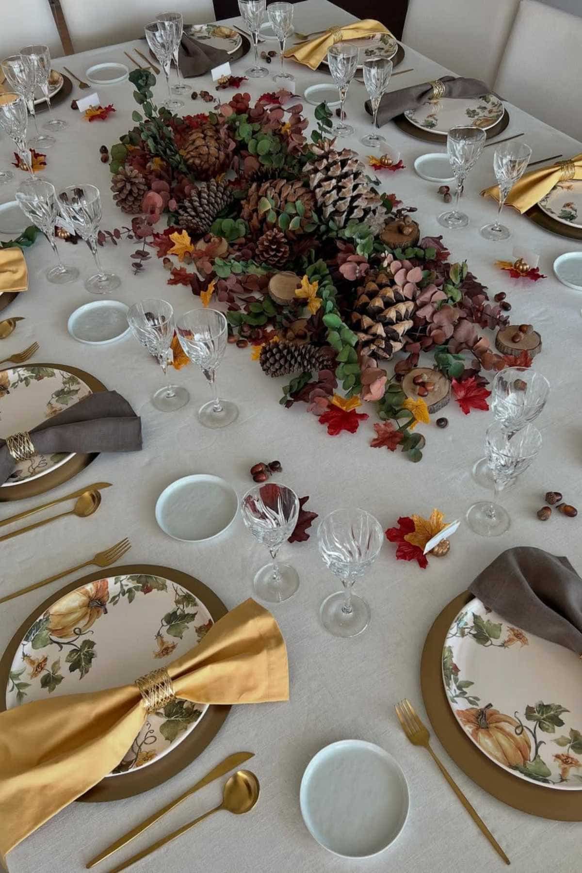 A dining table is set with autumn-themed plates, gold cutlery, folded napkins, crystal glasses, and a centerpiece of pinecones, leaves, and greenery.