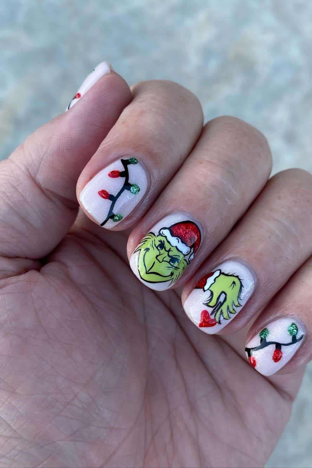 A hand with festive nail art featuring Christmas lights and illustrations of the Grinch's face and hand wearing a Santa hat.