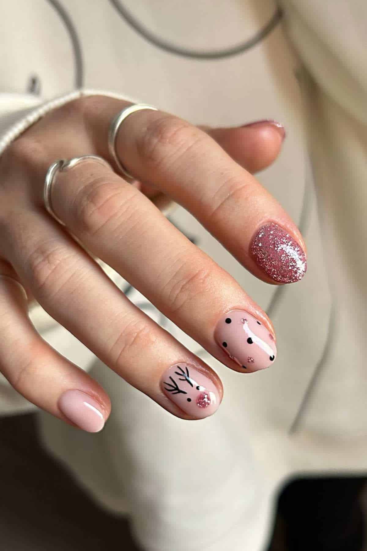 A hand with pink manicured nails, featuring a glitter accent nail and reindeer and polka dot designs on two nails.