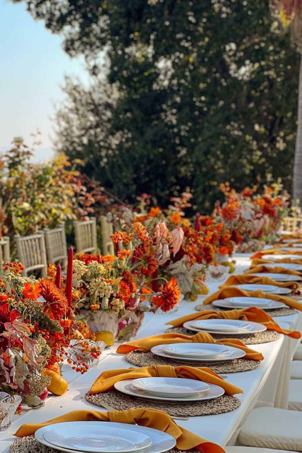 A long outdoor dining table is set with white plates, woven placemats, mustard yellow napkins, and autumn floral arrangements with red and orange flowers.